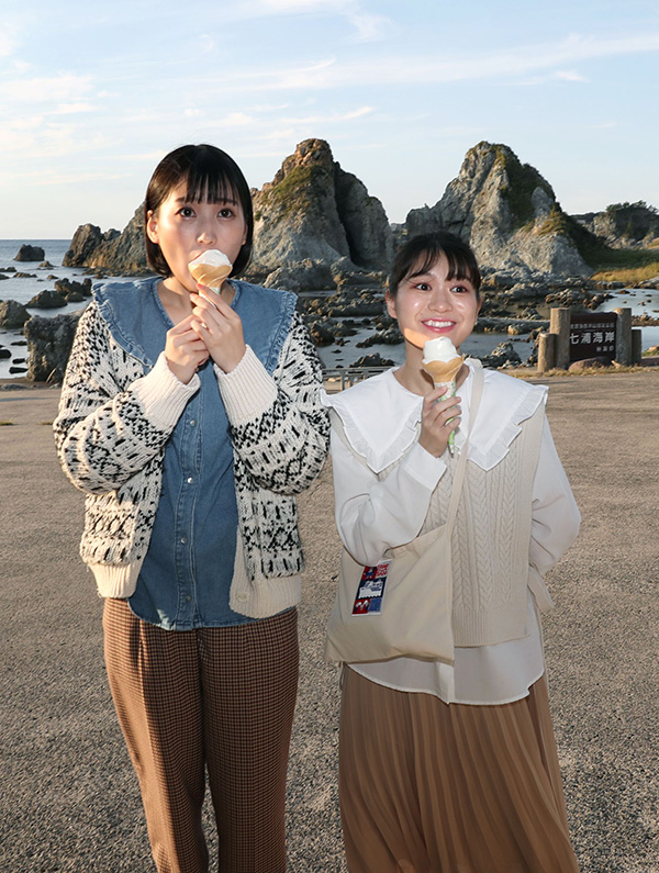 七浦海岸の名勝・夫婦岩をバックに、ミネラル豊富な佐渡の塩を使って作られたソフトクリームを頰張る宇野友恵さん（右）と横山実郁さん＝佐渡市高瀬